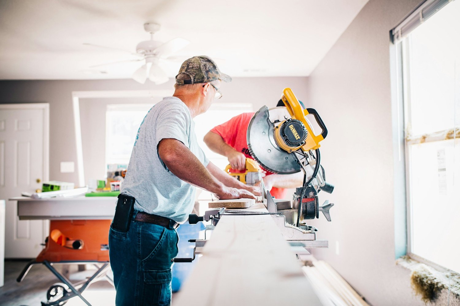 Two people on a job, cutting trim at a miter saw in an unfinished room.