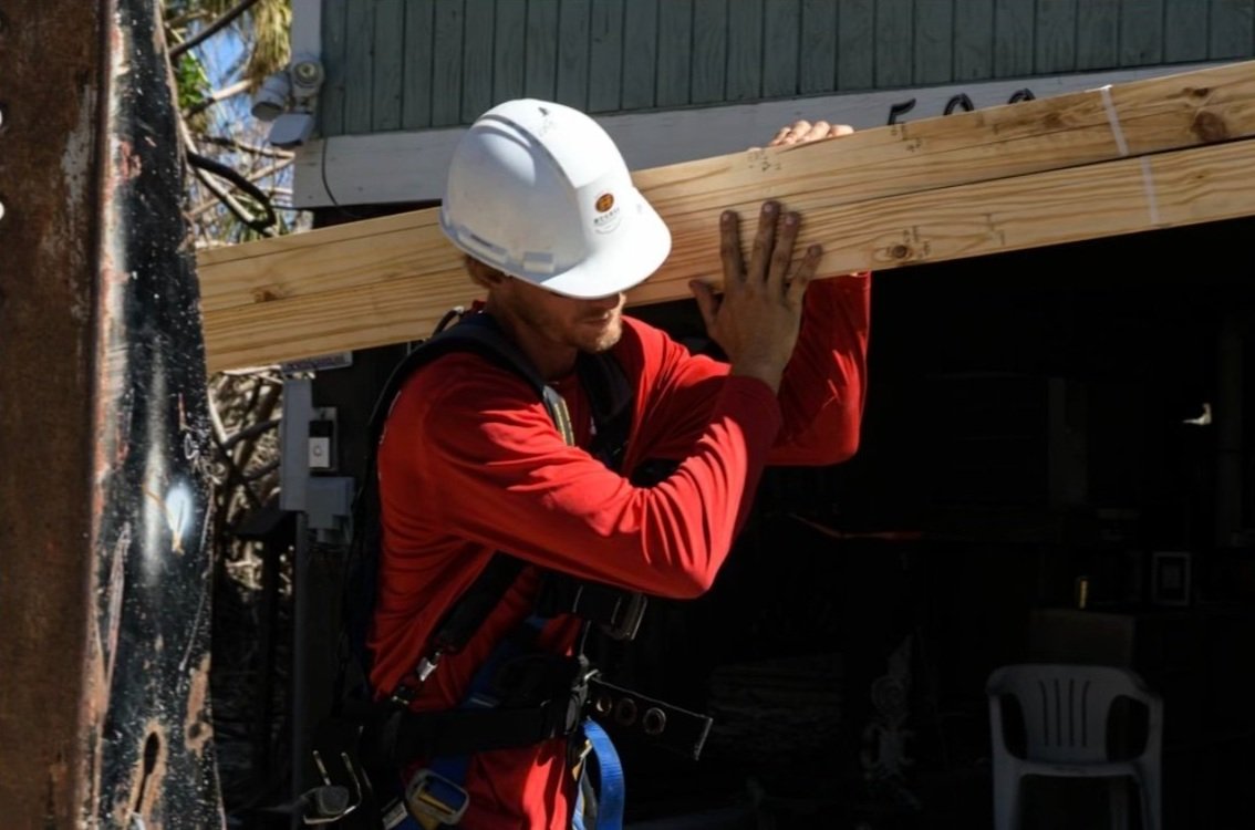 Brandon carrying dimensional lumber on a storm-repair job, hard hat and harness on.